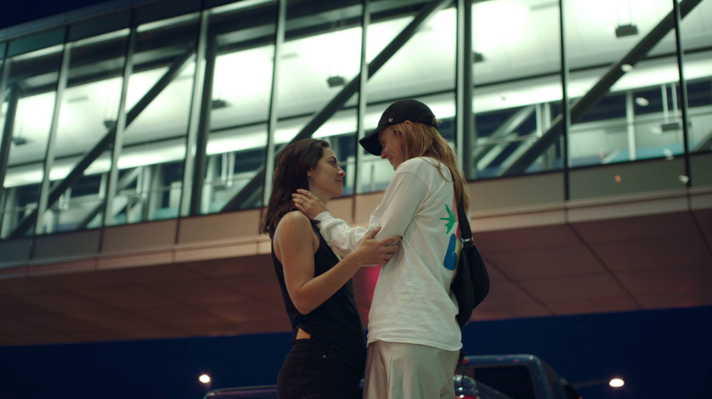 Feminin/Feminin (2014). Anne, who has long brown hair and black clothes, and Alex, who wears a baseball cap and a baggy white top, stand facing each other at an airport, smiling at each other. 