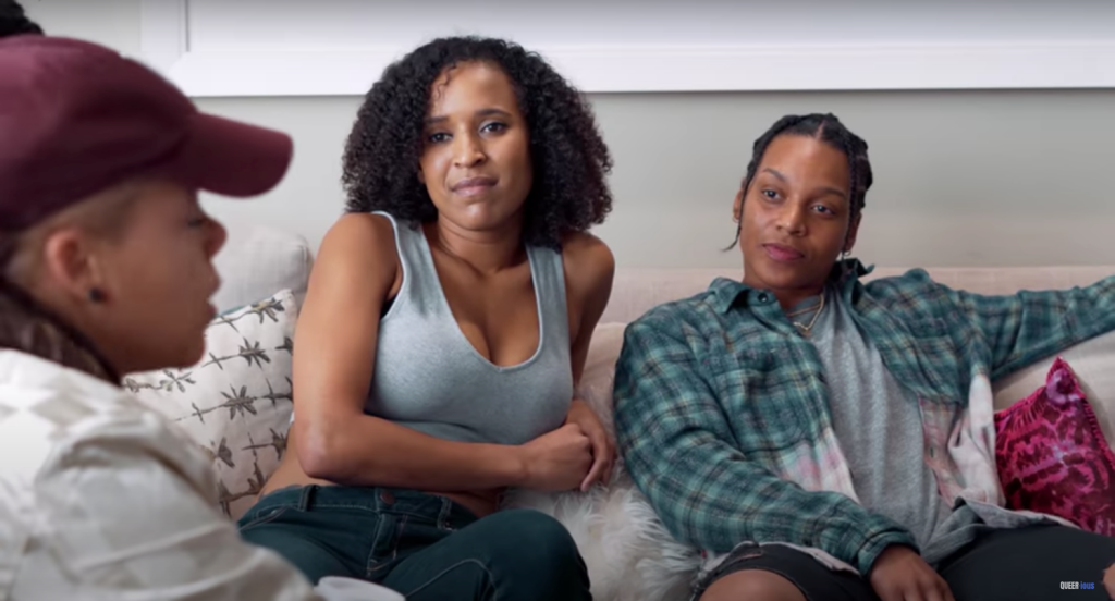 Queerious (2019). K'wame sits in the foreground wearing a baseball cap, talking to her friends Jazz and Bisa, who sit on the couch opposite. Jazz has curly hair and is dressed in a crop top, Bisa has braids and wears a blue flannel shirt.