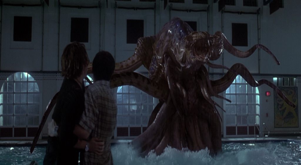 A still from 'The Faculty' Two men have their backs facing the camera in a school swimming pool room, they are holding each other at the shock of seeing a huge, fleshy, tentacled alien emerging from the pool.