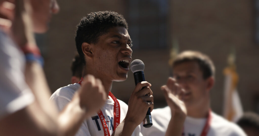 A still from documentary 'Boys State'. A teenage African American boy is shown in close-up speaking into a microphone at a mock political rally. Hands and arms of crowd members are blurred in the foreground and background. 