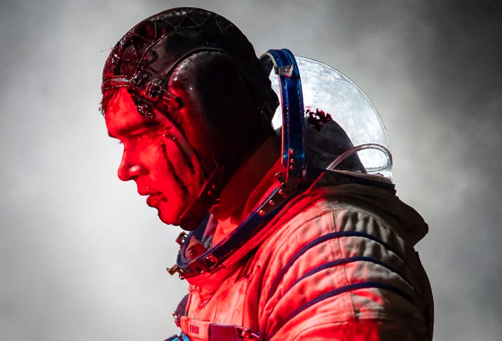 A still from 'Sputnik'. A man in a spacesuit is shown in a close-up profile view wearing his full astronaut gear, his helmet is tilted back and his head is covered in blood. The image is lit with blue and red lighting.