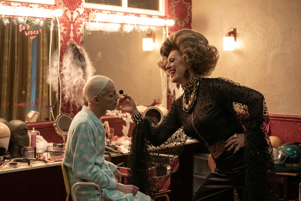 A still from 'Everybody's Talking About Jamie'. Jamie (Max Garwood) is say in a very cluttered dressing room, wearing a fluffy blue robe covered in white love hearts, his platinum hair concealed in a wig cap, he is getting made up by Loco Chanelle (Richard E. Grant), an ageing but glamorous drag queen.