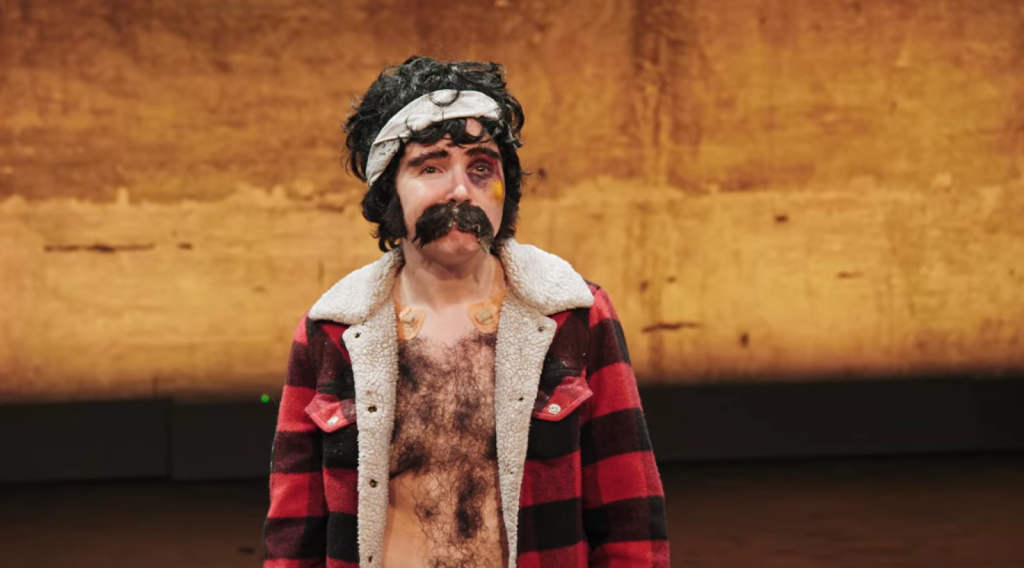 A half-body still depicting Nate at the beginning of the special, with his trademark white fleece-lined black and red lumberjack jacket open to expose his glued-on chest hair. Besides his angry yellow-purple blackeye (visible on his left eye), his prominent moustache, bushy eyebrows and unruly hair underneath a white bandana are visible. He’s standing on stage looking out over his audience outside of the frame. There’s something nonchalant and simultaneously tiresome about his facial expression, a sadness in his glistening eyes. 