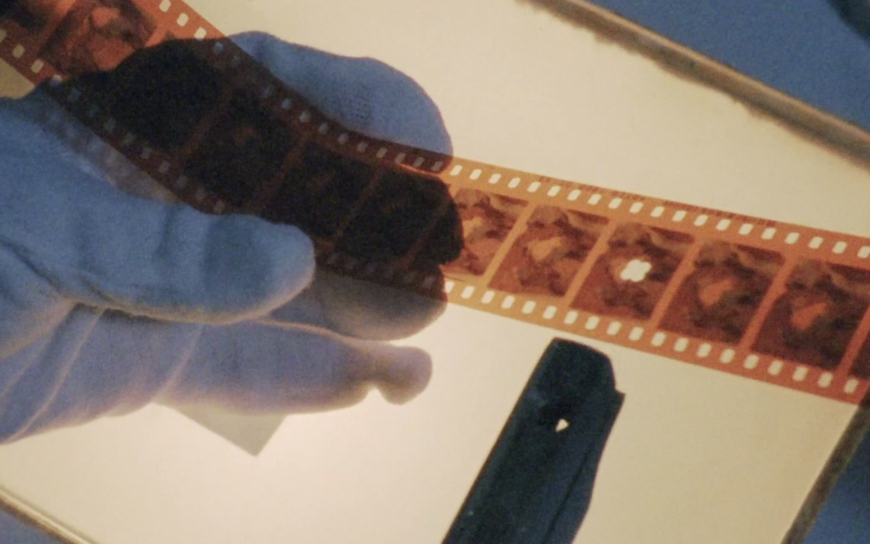 A still from Inventario Churubusco. Set in a film lab, this is a close up of a white gloved hand holding a strip of film against a lamp.